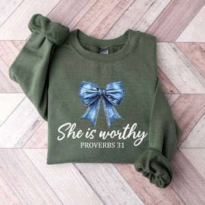 Może przedstawiać: Zielona bluza z niebieską kokardą z dżinsu i nadrukiem "She is worthy Proverbs 31" z przodu.