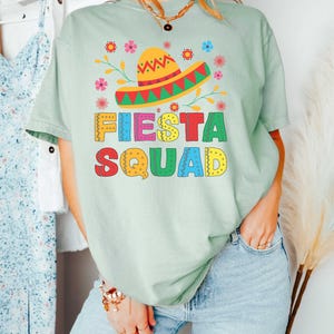 May include: A light green t-shirt with a colorful graphic of a sombrero and flowers. The text "FIESTA SQUAD" is written in colorful letters.