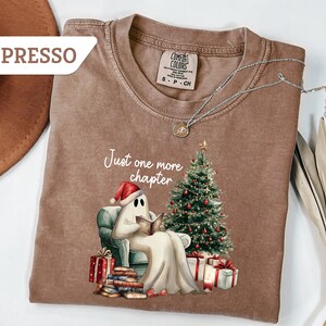 May include: Espresso-colored t-shirt featuring a ghost reading a book in front of a Christmas tree. The ghost wears a Santa hat and is surrounded by wrapped gifts. The text "Just one more chapter" is above the ghost.