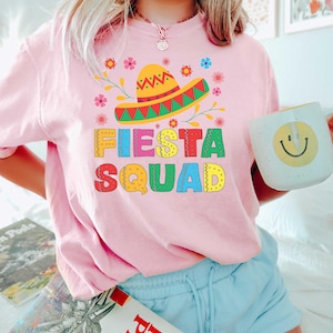 May include: Pink t-shirt with a colorful graphic of a sombrero and the text "Fiesta Squad".