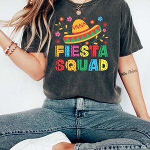 May include: A dark gray t-shirt with a colorful graphic of a sombrero, flowers, and the text "Fiesta Squad".