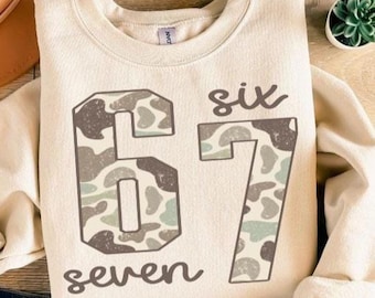 May include: Cream-colored sweatshirt with the number "67" in a camouflage pattern. The word "six" is above the "6" and the word "seven" is below the "7". The sweatshirt is folded and has a casual, relaxed style.