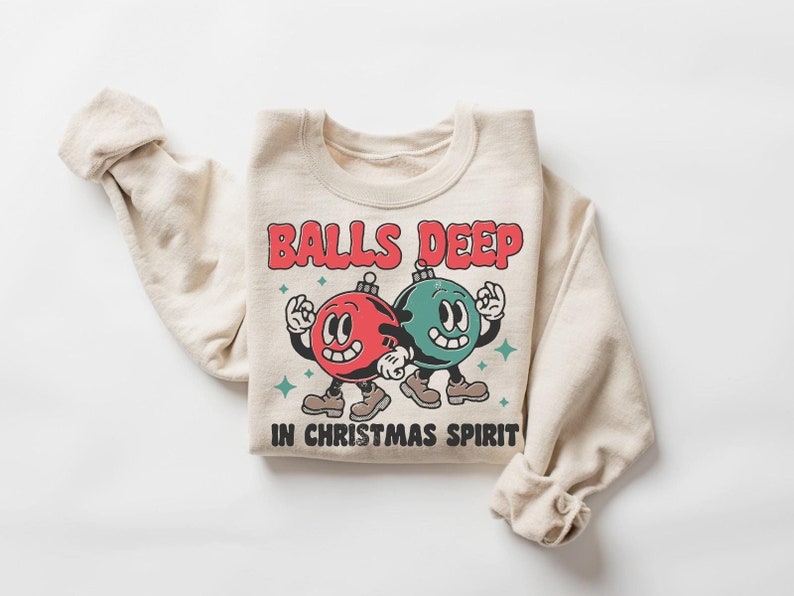 Peut inclure: Un sweat-shirt beige avec un motif de No&euml;l. Le motif repr&eacute;sente deux boules de No&euml;l dessin&eacute;es avec des visages, l'une rouge et l'autre verte, avec le texte "Balls Deep in Christmas Spirit".