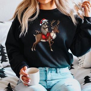 May include: A black sweatshirt with a Christmas-themed graphic of a raccoon wearing a Santa hat riding a reindeer. The raccoon is holding the reindeer's reins.
