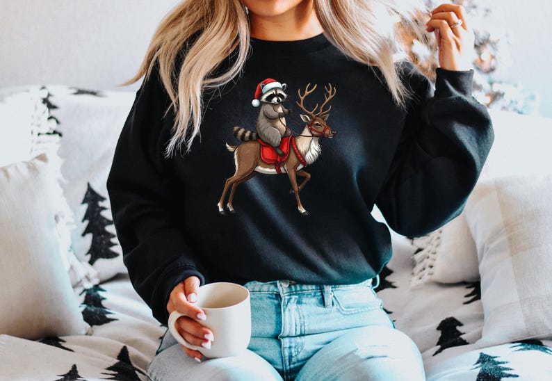 May include: A black sweatshirt with a Christmas-themed graphic of a raccoon wearing a Santa hat riding a reindeer. The raccoon is holding the reindeer's reins.
