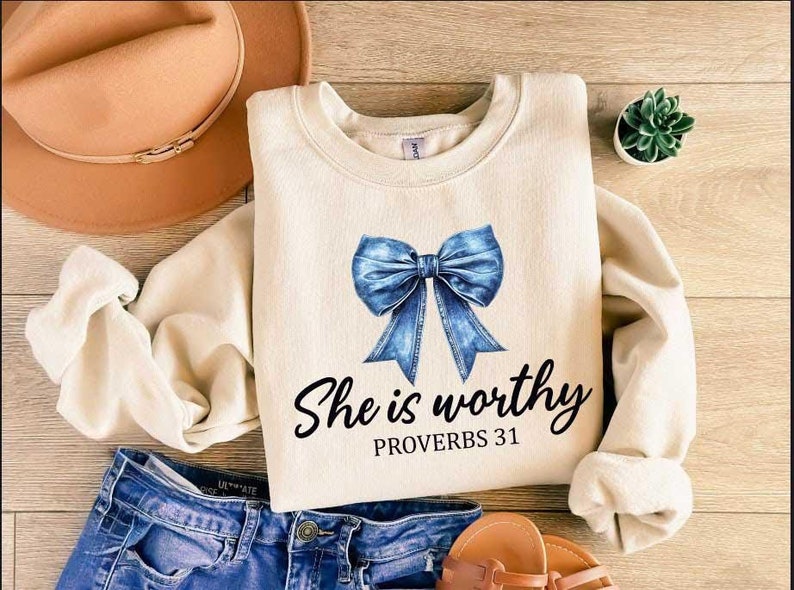 Może przedstawiać: Kremowa bluza z niebieską kokardą z dżinsu i nadrukiem "She is worthy Proverbs 31" z przodu.