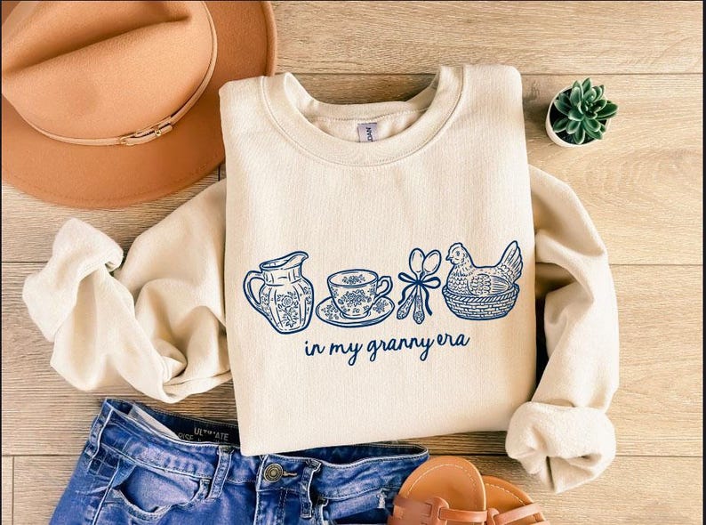 Puede incluir: Sudadera color crema con un dise&ntilde;o floral azul y el texto "in my granny era". El dise&ntilde;o incluye una jarra, una taza de t&eacute;, utensilios y una gallina en una cesta. Un sombrero marr&oacute;n y unos vaqueros azules tambi&eacute;n est&aacute;n en la imagen.