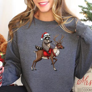 May include: A gray sweatshirt with a Christmas-themed design featuring a raccoon wearing a Santa hat riding a reindeer. The design is centered on the sweatshirt.