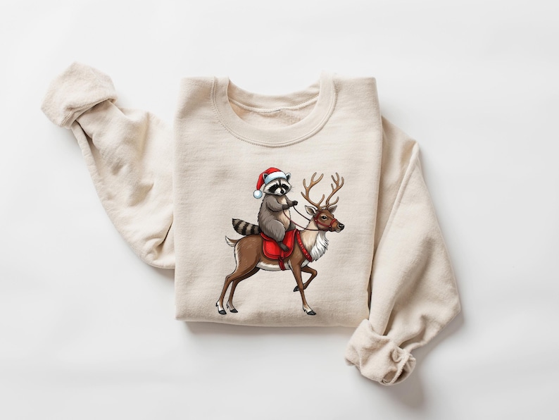 May include: A beige sweatshirt with a graphic of a raccoon wearing a Santa hat riding a reindeer.