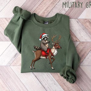 May include: A green Christmas sweatshirt with a raccoon wearing a Santa hat riding a reindeer. The sweatshirt has the text "MILITARY GREEN" printed on the top right corner.