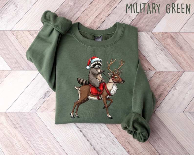 May include: A green Christmas sweatshirt with a raccoon wearing a Santa hat riding a reindeer. The sweatshirt has the text "MILITARY GREEN" printed on the top right corner.