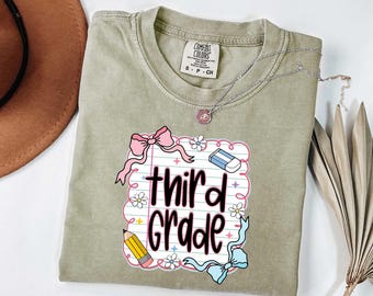 May include: A sage green t-shirt with a graphic that says "third grade" in black script. The graphic includes a pink bow, pencil, eraser, and flowers. A silver necklace with a small pendant is on the shirt.