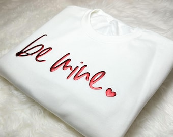 May include: White sweatshirt with the phrase "be mine" in red script with a small red heart.