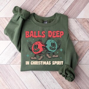 Peut inclure: Sweat-shirt vert avec un motif sur le th&egrave;me de No&euml;l. Le motif repr&eacute;sente deux ornements de No&euml;l en dessin anim&eacute; avec des visages, un rouge et un vert, et le texte "Balls Deep in Christmas Spirit".