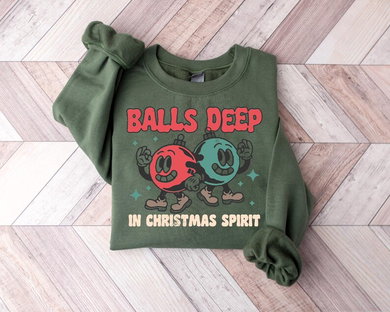 Peut inclure: Sweat-shirt vert avec un motif sur le th&egrave;me de No&euml;l. Le motif repr&eacute;sente deux ornements de No&euml;l en dessin anim&eacute; avec des visages, un rouge et un vert, et le texte "Balls Deep in Christmas Spirit".