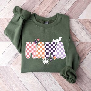 May include: A green sweatshirt with a checkered "MAMA" graphic in orange, pink, purple, and white. The graphic also includes a spider, a bat, and stars.