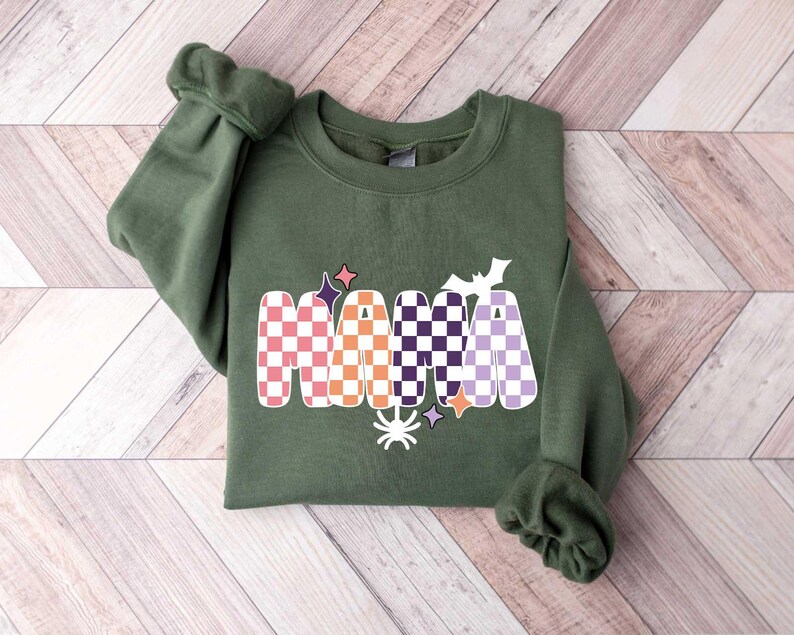 May include: A green sweatshirt with a checkered "MAMA" graphic in orange, pink, purple, and white. The graphic also includes a spider, a bat, and stars.