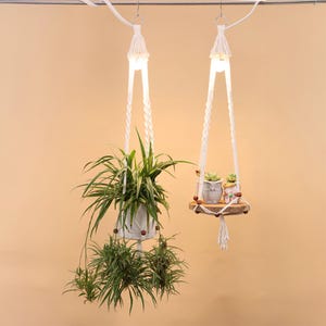 Pode incluir: Duas suspensões de plantas em macramé branco com plantas. Uma contém um vaso branco com uma planta verde, a outra uma pequena prateleira de madeira com duas pequenas suculentas em vasos. As suspensões são suspensas de uma barra de metal.