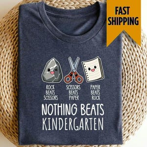 May include: A navy blue t-shirt featuring a Rock, Paper, Scissors design with the text "Nothing Beats Kindergarten". The design includes cartoon illustrations of a rock, scissors, and paper, with the corresponding text below each.