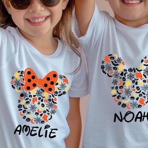May include: White t-shirts with a Halloween-themed Mickey Mouse design. The design features a bow with orange polka dots and a collection of Halloween icons. The names "Amelie", "Noah", and "Mia" are printed below the designs.