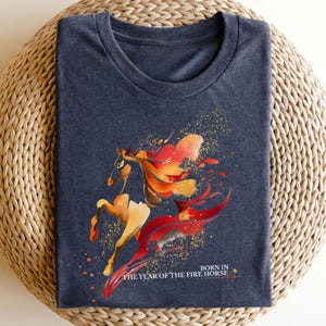 Pu&ograve; includere: T-shirt blu navy con un design stilizzato di cavallo nei toni dell'arancione, rosso e oro. Il design include un effetto splash e il testo "BORN IN THE YEAR OF THE FIRE HORSE".
