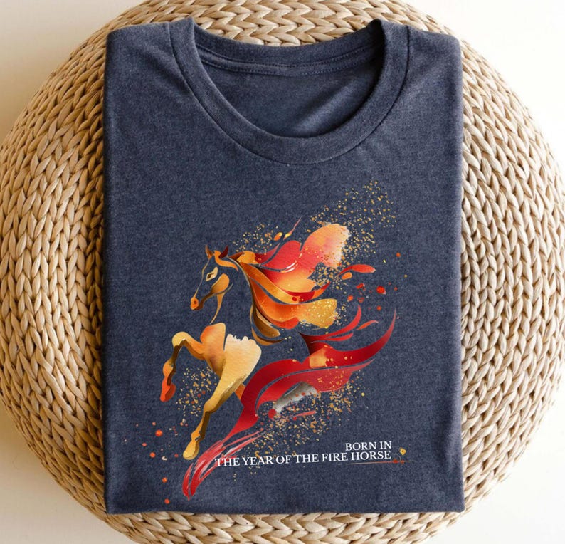 Pu&ograve; includere: T-shirt blu navy con un design stilizzato di cavallo nei toni dell'arancione, rosso e oro. Il design include un effetto splash e il testo "BORN IN THE YEAR OF THE FIRE HORSE".