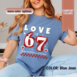 May include: Blue jean colored t-shirt with the text "LOVE IS 67" in white and red. The number 67 is in a bold, outlined font. The shirt also features the phrase "COZY MOMENTS WITH YOU" and heart graphics.