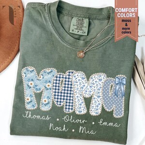 Pu&ograve; includere: T-shirt verde oliva con la scritta "MOM" in un design patchwork. Le lettere sono decorate con motivi floreali e a quadri, e un fiocco blu. I nomi sono stampati sotto la parola "MOM". &Egrave; visibile una collana con un ciondolo.