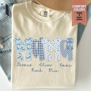 Pu&ograve; includere: T-shirt color crema con la scritta "MAMA" in motivi floreali, a quadri e a pois blu. I nomi Thomas, Oliver, Emma, Noah e Mia sono stampati sotto. Un fiocco blu &egrave; sull'ultima lettera. La maglietta &egrave; piegata.