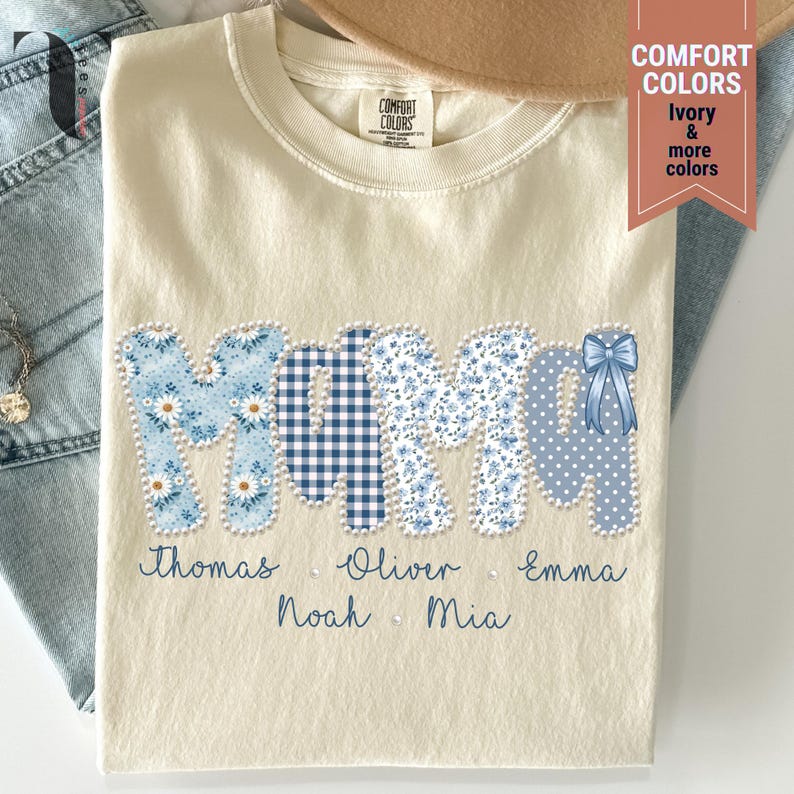 Pu&ograve; includere: T-shirt color crema con la scritta "MAMA" in motivi floreali, a quadri e a pois blu. I nomi Thomas, Oliver, Emma, Noah e Mia sono stampati sotto. Un fiocco blu &egrave; sull'ultima lettera. La maglietta &egrave; piegata.
