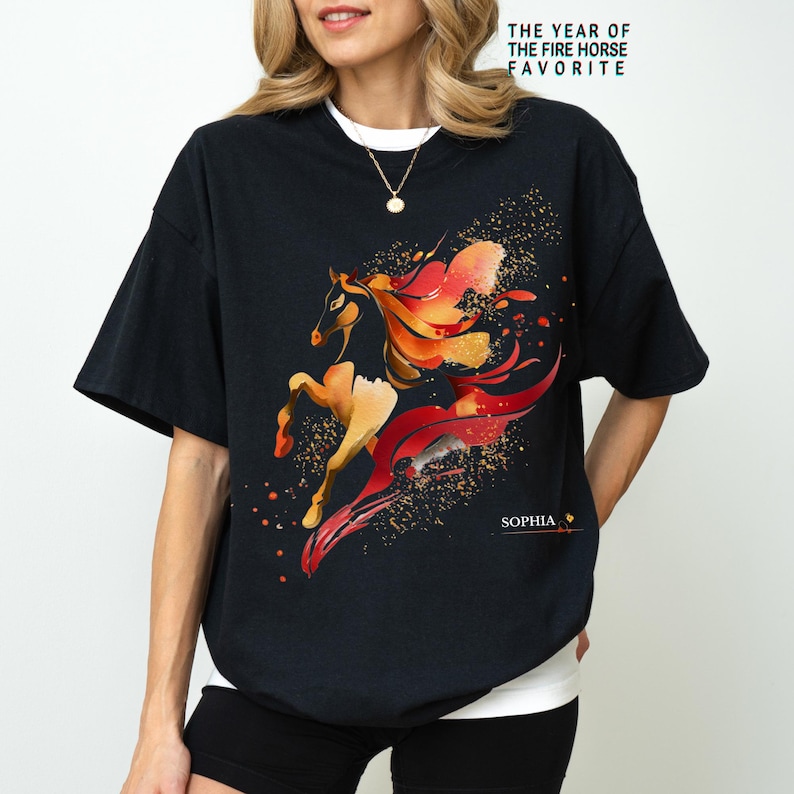 Pu&ograve; includere: T-shirt nera con un disegno ad acquerello di un cavallo nei toni dell'arancione, del rosso e dell'oro. Il testo "THE YEAR OF THE FIRE HORSE FAVORITE" &egrave; stampato in alto, con il nome "SOPHIA" in basso.