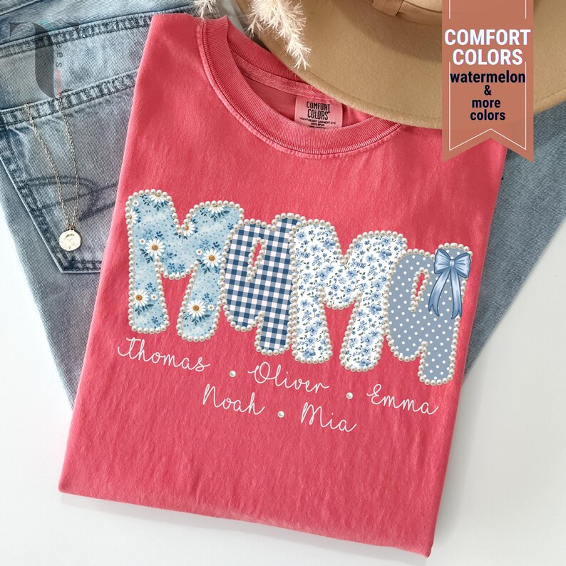 Pu&ograve; includere: Una maglietta color corallo con la scritta "MAMA" in lettere patchwork. Le lettere presentano motivi floreali, a quadretti e a pois. Sotto ci sono i nomi Thomas, Oliver, Noah, Mia ed Emma.