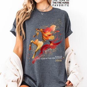 Pu&ograve; includere: T-shirt grigio scuro con un design stilizzato di cavallo nei toni del rosso, arancione e oro. Il testo "THE YEAR OF THE FIRE HORSE FAVORITE" e "BORN IN THE YEAR OF THE FIRE HORSE 1966" &egrave; stampato sulla maglietta.