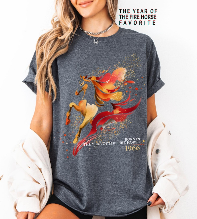 Pu&ograve; includere: T-shirt grigio scuro con un design stilizzato di cavallo nei toni del rosso, arancione e oro. Il testo "THE YEAR OF THE FIRE HORSE FAVORITE" e "BORN IN THE YEAR OF THE FIRE HORSE 1966" &egrave; stampato sulla maglietta.
