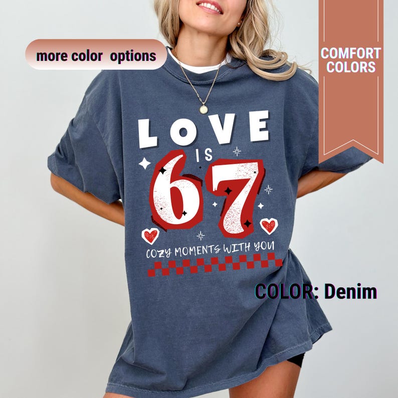 May include: Denim blue t-shirt with the text "LOVE IS 67" in white and red. The shirt has a relaxed fit and features a graphic with hearts and stars. The words "COZY MOMENTS WITH YOU" are also printed on the shirt.