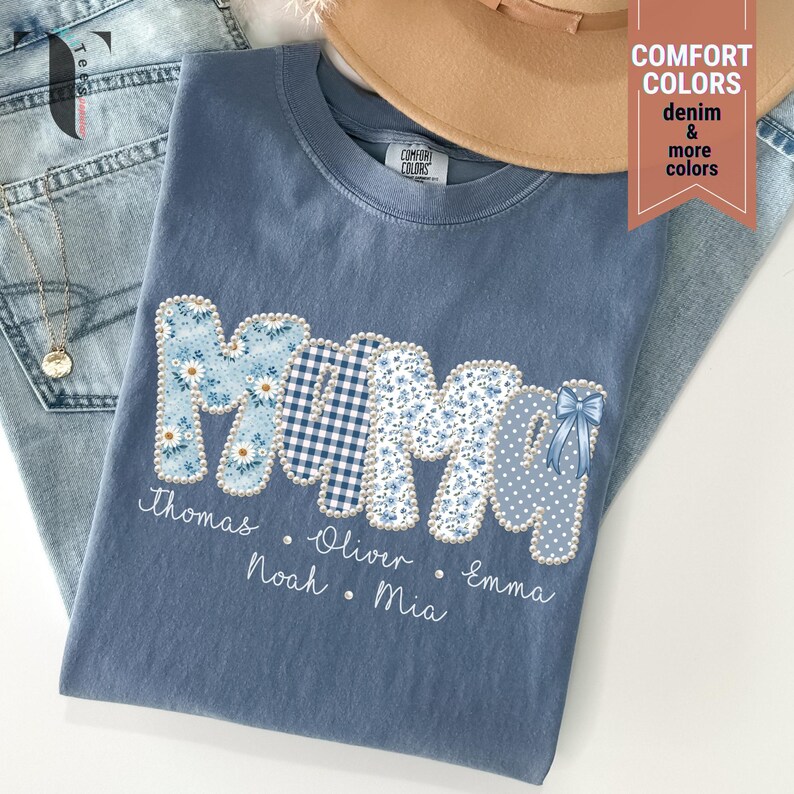 Pu&ograve; includere: Una maglietta blu Comfort Colors con la scritta "MAMA" in un design patchwork. Le lettere presentano motivi floreali, a quadri e a pois. Sotto la parola "MAMA" ci sono i nomi Thomas, Oliver, Noah, Emma e Mia. Un cappello color cachi e jeans sono sullo sfondo.