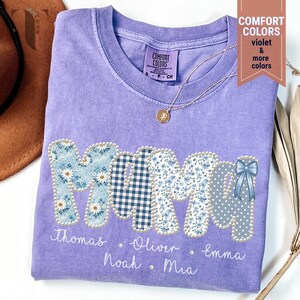 Pu&ograve; includere: Una maglietta Comfort Colors color lavanda con la scritta "MAMA" in un design patchwork. Le lettere sono decorate con motivi floreali, quadretti e pois. I nomi sono stampati sotto le lettere. Una collana d'oro &egrave; drappeggiata sulla maglietta.