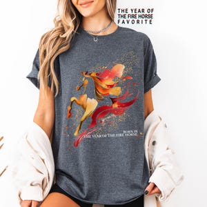 Pu&ograve; includere: T-shirt grigio scuro con un disegno ad acquerello di un cavallo nei toni dell'arancione e del giallo con accenti rossi. Il testo "THE YEAR OF THE FIRE HORSE FAVORITE" e "BORN IN THE YEAR OF THE FIRE HORSE" &egrave; stampato.
