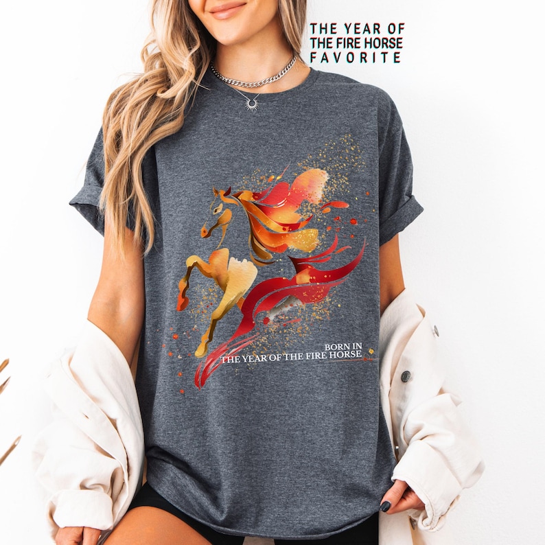 Pu&ograve; includere: T-shirt grigio scuro con un disegno ad acquerello di un cavallo nei toni dell'arancione e del giallo con accenti rossi. Il testo "THE YEAR OF THE FIRE HORSE FAVORITE" e "BORN IN THE YEAR OF THE FIRE HORSE" &egrave; stampato.