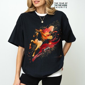Pu&ograve; includere: T-shirt nero con un disegno ad acquerello di un cavallo nei toni dell'arancione, del rosso e dell'oro. Il cavallo &egrave; raffigurato in movimento con criniera e coda fluenti. Il testo "THE YEAR OF THE FIRE HORSE FAVORITE" &egrave; stampato in alto.