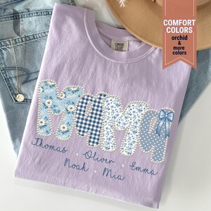 Pu&ograve; includere: Una maglietta lilla Comfort Colors con la scritta "MOM" in un design patchwork. Le lettere sono decorate con motivi floreali, a quadri e a pois. I nomi sono ricamati sotto la parola "MOM".