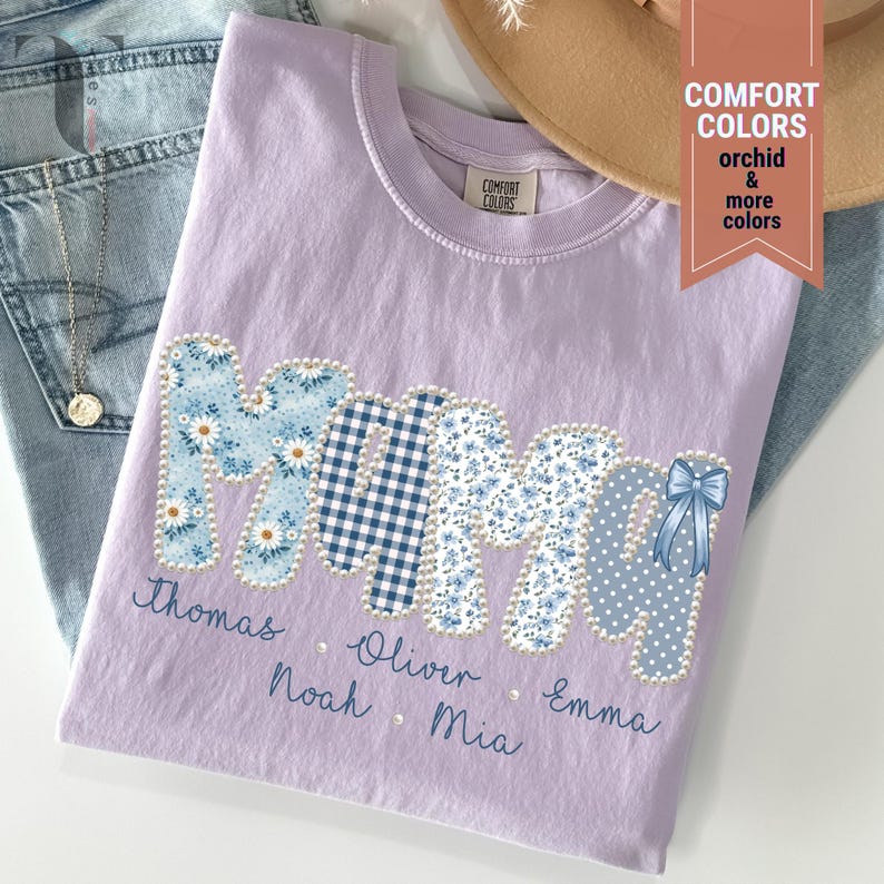 Pu&ograve; includere: Una maglietta lilla Comfort Colors con la scritta "MOM" in un design patchwork. Le lettere sono decorate con motivi floreali, a quadri e a pois. I nomi sono ricamati sotto la parola "MOM".