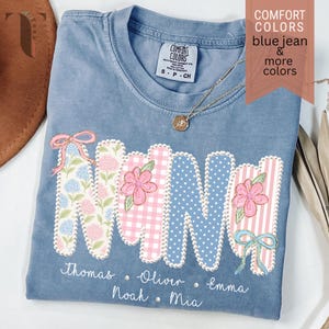 May include: Blue jean Comfort Colors t-shirt with the word "NINI" in a floral design. The shirt includes names below the word and the text "COMFORT COLORS blue jean & more colors". A gold necklace is visible.