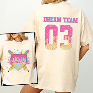 May include: Cream-colored t-shirt with a graphic on the front and back. The front graphic features pencils, a pink ice cream design, and the text "Third Grade Drip." The back displays "DREAM TEAM" above the number "03" in a pink and waffle-like design.