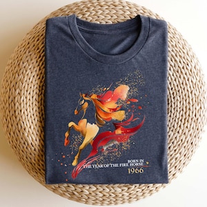 Pu&ograve; includere: T-shirt blu navy con un'illustrazione stilizzata di un cavallo nei toni dell'arancione, rosso e oro. Il testo "BORN IN THE YEAR OF THE FIRE HORSE 1966" &egrave; stampato sotto. Il cavallo ha una criniera e una coda fluenti.