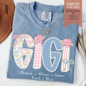 May include: A blue jean Comfort Colors t-shirt with the name "GIGI" in a floral and patterned design. The shirt includes the names Thomas, Oliver, Emma, Noah, and Mia. A gold necklace is draped across the shirt.