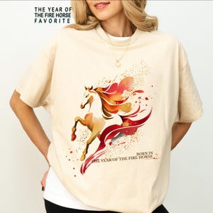 Pu&ograve; includere: T-shirt color crema con un'illustrazione ad acquerello di un cavallo con accenti rossi e arancioni. Il testo "THE YEAR OF THE FIRE HORSE FAVORITE" e "BORN IN THE YEAR OF THE FIRE HORSE" sono stampati sulla maglietta.