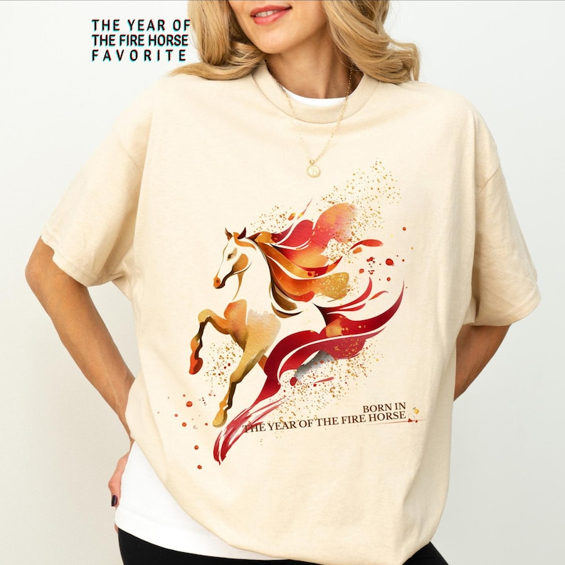 Pu&ograve; includere: T-shirt color crema con un'illustrazione ad acquerello di un cavallo con accenti rossi e arancioni. Il testo "THE YEAR OF THE FIRE HORSE FAVORITE" e "BORN IN THE YEAR OF THE FIRE HORSE" sono stampati sulla maglietta.