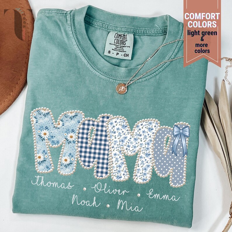 Pu&ograve; includere: Una maglietta Comfort Colors verde chiaro con la scritta "MAMA" in un design patchwork. Le lettere sono decorate con motivi floreali e gingham, e un fiocco blu. I nomi sono stampati sotto.