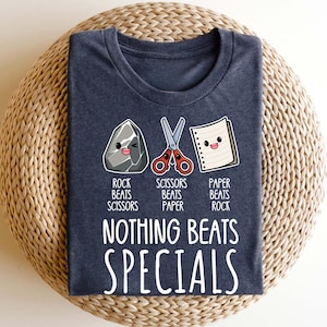 May include: A dark heather gray t-shirt featuring a graphic of a rock, scissors, and a piece of paper with faces. The text reads: ROCK BEATS SCISSORS, SCISSORS BEATS PAPER, PAPER BEATS ROCK, and NOTHING BEATS SPECIALS in white lettering across the chest.